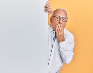 Senior caucasian man holding blank empty banner covering mouth with hand, shocked and afraid for mistake. surprised expression